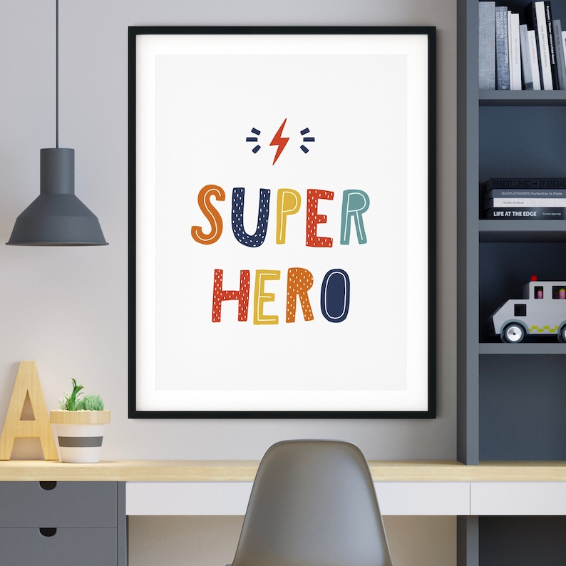 以下が含まれることがあります： 白い背景にカラフルな文字で「SUPER HERO」と書かれた黒い額縁のプリント。文字の上に赤い稲妻が描かれている。