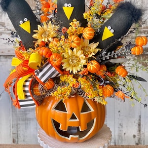May include: A Halloween centerpiece featuring a carved orange pumpkin vase with a smiling face. The vase holds a floral arrangement with black crow figures, sunflowers, and miniature pumpkins. The arrangement is accented with ribbons.