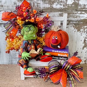 May include: A green Frankenstein-inspired plush figure wearing glasses and reading a book, seated on a white bench with a stack of books titled Frankenstein, Dracula, and Sleepy Hollow, topped with a plush pumpkin and orange bows.