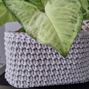 May include: A gray crocheted plant basket with a green plant inside. The basket is made of thick rope and has a textured surface. The text "MK Cord Store" is visible in the lower left corner of the image.