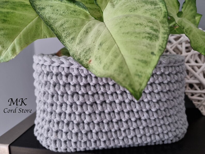 May include: A gray crocheted plant basket with a green plant inside. The basket is made of thick rope and has a textured surface. The text "MK Cord Store" is visible in the lower left corner of the image.