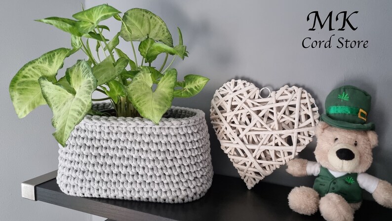 May include: A gray crocheted planter with a green leafy plant inside. The planter is on a shelf with a white wicker heart and a teddy bear wearing a green hat.