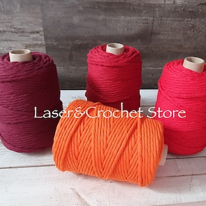 Peut inclure: Trois bobines de corde en coton de couleur rouge, orange et bordeaux. Les bobines sont disposées sur une surface en bois blanc.  Laser & Crochet Store est imprimé sur la surface.