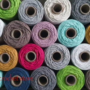 Op de afbeelding: Een verzameling van 20 spoelen katoenen koord in verschillende kleuren, waaronder wit, grijs, groen, blauw, roze en geel. De spoelen zijn gerangschikt in een rasterpatroon.