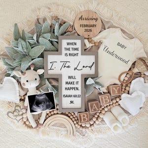 May include: A baby announcement featuring a cream-colored onesie with "BABY Underwood" printed on it, surrounded by wooden blocks spelling "BABY", a crochet giraffe, and a wooden cross with a Bible verse. A wooden sign says "Arriving February 2026".