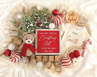 Könnte beinhalten: Eine weihnachtliche Babyankündigung. Ein rot gerahmtes Schild liest "THE MOST SPECIAL Christmas Gift IS ON THE WAY! APRIL 2026". Ein Teddybär, Babykleidung und Ornamente sind auf einer cremefarbenen Decke angeordnet.