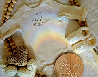 Puede incluir: Un body de bebé beige con un diseño de arcoíris y el texto "BABY Blair". Un letrero de madera con el texto "Arriving 10.2025" y un símbolo de corazón. Un juguete de corazón tejido a crochet y un collar de cuentas de madera.