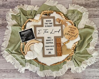 May include: A pregnancy announcement featuring a green blanket with white fringe, a wooden cross with text, a sonogram, and wooden blocks spelling "BABY." Decorative elements include a wooden rainbow, hearts, and a wooden disc.