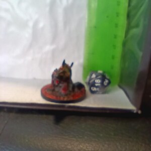 May include: A miniature painted figure of a creature with a red and black body, blue eyes, and a black base. The figure is standing on a black and red base with a white rim. A blue and white dice is in the background.