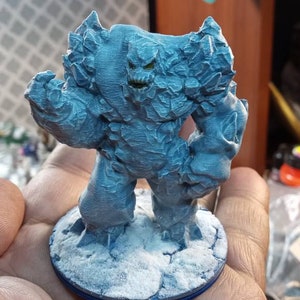 May include: A blue and white miniature figure of a rock golem, standing on a snowy base. The figure is detailed with textured rock formations and a menacing expression.