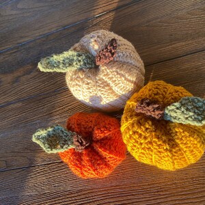 Puede incluir: Tres calabazas de crochet en diferentes colores: beige, naranja y amarillo. Cada calabaza tiene un tallo de crochet verde.