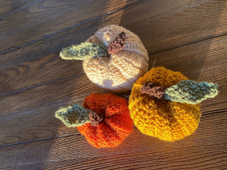 Puede incluir: Tres calabazas de crochet en diferentes colores: beige, naranja y amarillo. Cada calabaza tiene un tallo de crochet verde.