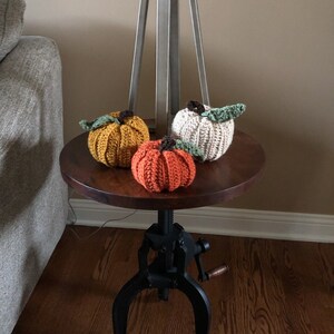 Puede incluir: Tres calabazas de crochet en colores naranja, amarillo y beige, sentadas en una mesa de madera con una base de metal negro.