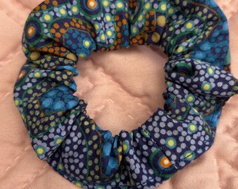 May include: A blue, green, and orange patterned scrunchie with a white background. The scrunchie is made of fabric and has a gathered look.