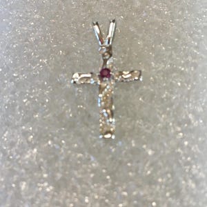 May include: A silver cross pendant with a small, round, red gemstone at the center. The cross has a textured surface and a loop at the top for a chain. The background is a soft, white, textured surface.