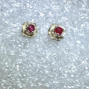 May include: A pair of stud earrings featuring a small, round, vibrant red gemstone set in the center of a textured, gold-toned metal flower-shaped setting. The earrings are designed with a post back for secure attachment to the earlobe.
