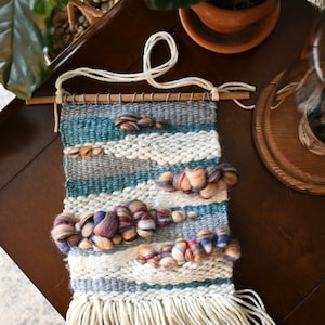 May include: A woven wall hanging with a white, gray, and teal color scheme. The hanging features a fringe at the bottom and a variety of colorful yarn balls woven into the design.