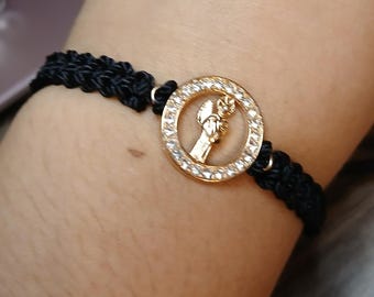 May include: Black braided bracelet with a gold-colored circular charm. The charm features a raised figure within a circle of clear, sparkling stones. The bracelet is secured with a sliding knot closure.