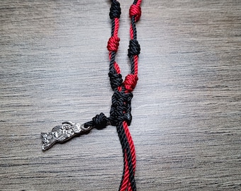 May include: A handmade rosary featuring red and black knotted cord. The rosary has a silver-toned metal pendant and two silver beads at the end. The cord is knotted to form the beads.