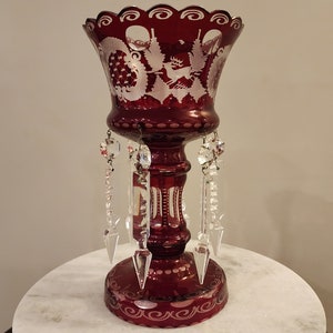 May include: A red glass vase with a white etched design of a deer and trees. The vase has a scalloped rim and is decorated with clear crystal prisms.