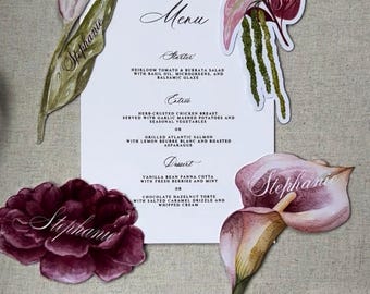 Op de afbeelding: Een witte rechthoekige menukaart met zwarte tekst met gerechten zoals tomaten-burratasalade, kruidenkip en vanille-panna cotta, omringd door aquarel bloemenuitsparingen met de naam Stephanie in sierlijk schrift.