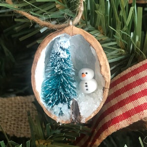 May include: A small, handmade Christmas bauble made from a walnut shell. The bauble features a miniature pine tree and a snowman inside the shell, with white glitter representing snow.