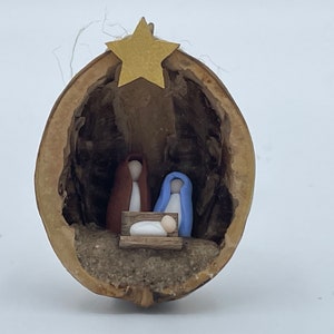 May include: A miniature nativity scene inside a hollowed-out walnut shell. The figures are made of clay and depict Mary, Joseph, and the baby Jesus. A gold star sits above the scene.
