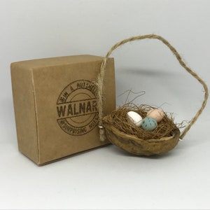 May include: A small, brown, wooden walnut shell with a twine handle, filled with straw and three miniature eggs. The eggs are white, pink, and blue with white speckles. The walnut shell is hanging from a brown cardboard box with a logo that reads "I'm a Nutshell, Walnar, Surprising Nuts".