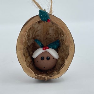 May include: A Christmas ornament made from a walnut shell with a small, brown, clay figure inside. The figure is wearing a white hat with red and green accents and has black eyes.