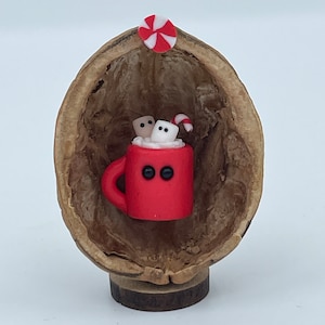 May include: A miniature red mug with two black eyes, filled with white foam, two marshmallows, and a candy cane, nestled inside a hollowed-out walnut shell. A small red and white peppermint candy is attached to the top of the walnut shell.