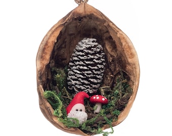 May include: A small, decorative ornament made from a halved walnut shell. Inside the shell, there is a miniature gnome, a small red mushroom, and a pine cone covered in white glitter.