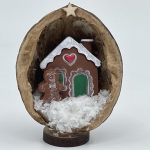 May include: A gingerbread house and gingerbread person figurine inside a hollowed-out walnut shell with white faux snow. A wooden star sits on top of the walnut shell.