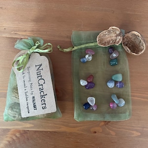May include: Two green organza bags with drawstring closures. One bag contains a small brown paper tag with the text "Nut Crackers Surprising Nuts by WALMART Crack to reveal a hidden surprise". The other bag contains a variety of small colourful stones.