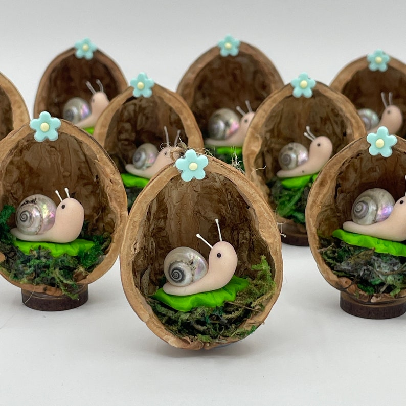 A miniature pale pink polymer clay snail body, with pearlescent real shell body and paper antennae and black eyes, sat on top of a bright green polymer clay leaf all inside a real half walnut shell. Image shows several items stood in a group.