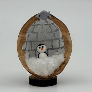 May include: A miniature diorama inside a brown walnut shell featuring a small black and white penguin standing on white faux snow. A translucent igloo structure with drawn grey brick lines sits behind the penguin, and a small grey fish is attached to the top.