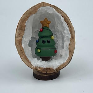 May include: A small green Christmas tree with colorful ornaments is inside a hollowed-out walnut shell. The walnut shell is sitting on a wooden base.