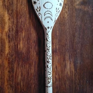 May include: Wooden spoon with a light wood finish, featuring a burned design of moon phases and floral accents. The handle is adorned with a repeating floral pattern. The text "Design 1" is visible at the bottom of the spoon.