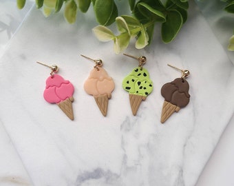 May include: Four pairs of ice cream cone earrings. The earrings are made of polymer clay and feature different ice cream flavors: pink, peach, green with chocolate chips, and brown. The earrings are attached to gold-colored hooks.