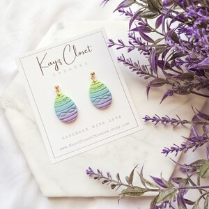 May include: A pair of pastel green, blue, and purple Easter egg-shaped earrings with gold hooks. The earrings are handmade and feature a textured design. The earrings are displayed on a white card with the text "Kayz Closet Creates" and a website address.