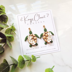 May include: Gold and green turtle earrings with white flower accents. The earrings are handmade and feature the text "Kayz Closet Creates" and "Handmade with Love" on the card they are displayed on.