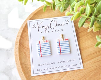 May include: A pair of gold stud earrings with a white paper notepad design. The notepad has blue lines and a red line at the top. The notepad has a red "A+" grade on it. The earrings are on a white card with the text "Kayz Closet Creates" and "Handmade with Love Kayzclosetcreates.etsy.com".
