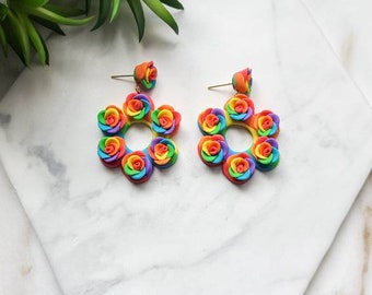 May include: A pair of rainbow-colored rose earrings with gold hooks. Each earring features a circle of six roses, with each rose having a different color of the rainbow.