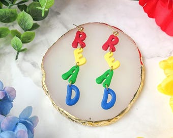 May include: A pair of dangle earrings with the word "READ" spelled out in colorful letters. The letters are red, yellow, green, and blue.