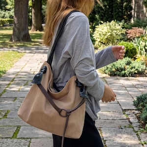 May include: A beige hobo-style shoulder bag featuring black leather-like knotted accents at the top corners and a thin dark brown strap that loops through a circular metal ring on the front, creating a draped and casual silhouette.
