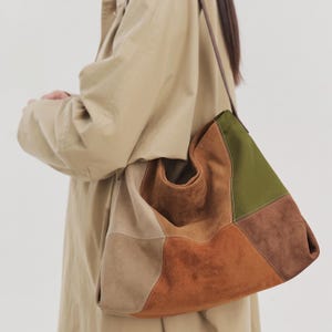 May include: A slouchy hobo bag crafted from panels of suede in various earth tones, including olive green, dark brown, rust orange, and beige. The bag features visible white stitching along the seams and a thin, dark brown leather shoulder strap.