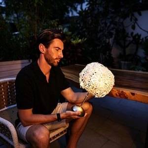 May include: A table lamp featuring a spherical shade constructed from numerous white, daisy-shaped cutouts. The lamp has a slender metallic stem and a smooth, rounded silver base, emitting a warm, soft glow from within the floral-patterned structure.