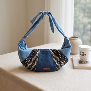 May include: A crescent-shaped hobo bag featuring a light blue top section with a knotted handle, a dark blue center panel with a quilted texture, and black side panels adorned with a white floral pattern. A small brown rectangular label reads Star-flying.