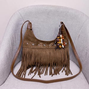 May include: A brown suede hobo bag featuring a long fringe trim along the bottom, a row of silver-toned star-shaped studs across the center, and a decorative charm made of various brown and cream-colored wooden beads attached to the strap.