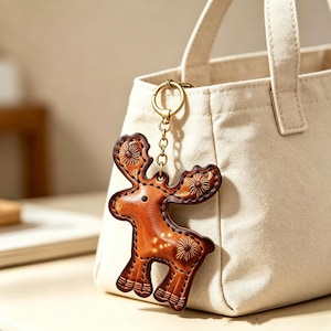 May include: A tan leather keychain shaped like a stylized moose with embossed floral patterns on the antlers and body, featuring dark brown stitched edges and a gold-toned metal ring attached to a cream-colored canvas tote bag with two handles.