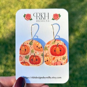 May include: Dangle earrings featuring a beige background with a repeating pattern of orange pumpkins with smiling faces and small green leaves. The earrings are displayed on a white card labeled RKH DESIGNS with rose icons.